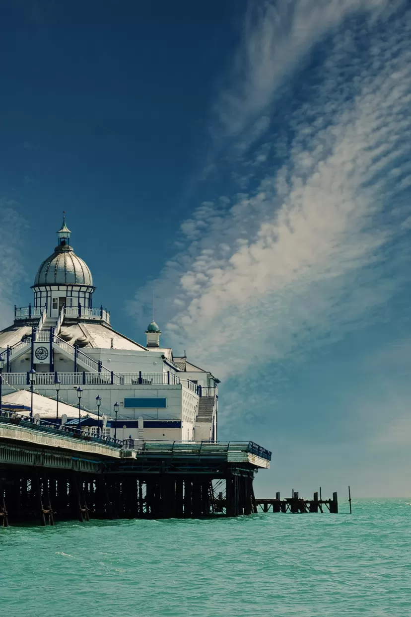 A stunning photo of Eastbourne pier taken on a sunny day. The photo is taken from low down, on the beach probably, so that the white pier looks particularly imposing. The sky is blue with light streaks of cloud and the sea is an inviting pale jade colour.