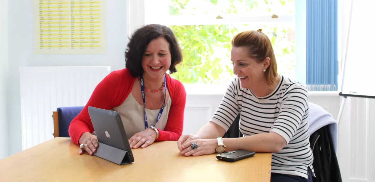 A student and teacher laugh together as they look at a tablet device in what looks to be a one to one classroom at the English language centre in Chester. The one to one component is part of the 25 plus combination course at the school.