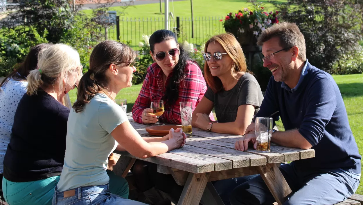 Students on the 25 plus English course in Chester take time out to have a few drinks around an outdoor picnic table with their teacher. The setting looks to be an English pub garden and the picnic table is placed on a green lawn.