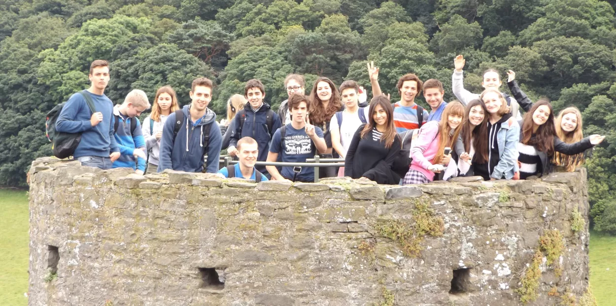 A photo of a large group of ELC English students all standing at the very top of a round tower at Conwy castle in North Wales. The tower is very old and is made of stone and the photograph has been taken from another tower or a position equally high up on the castle walls. In the background we can see lots of trees in a dense woodland.