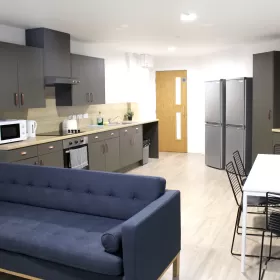 A photo of a kitchen in a student residence. This looks quite spacious and you can see a large double fridge freezer in the background and a blue sofa in the foreground.