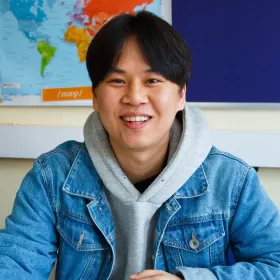 South Korea's Young Huon Song, a student on the General English programme at ELC Chester is smiling openly at the camera, whilst sitting in one of the classrooms in the Chester English school. He's wearing a denim jacket over a light grey hoodie. He looks very relaxed.
