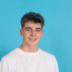 A teenage student looking into the camera lens, wearing a plan white T-shirt on a plain blue background.
