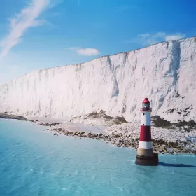 A professional photo of the red and white striped lighthouse just off the East Sussex coast by Eastbourne. Whilst the lighthouse is impressive, so too are the white chalk cliffs which take up a third of the photo as they disappear off into the distance to the left of the photo. The rest of the photo is of blue sky and blue sea.