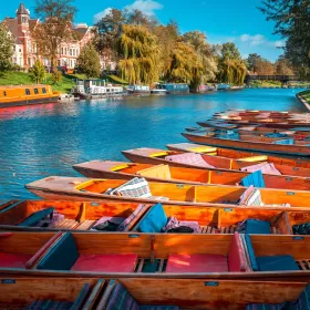 A colourful photo of punts, traditional wooden boats, on the River Cam in Cambridge. The water looks particularly blue, as does the sky, and whilst a number of boats are tied up on one side of the river, you can see a beautiful red brick building and several willow trees in the distance on the opposite bank.
