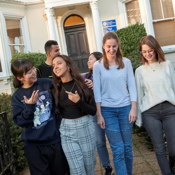 A group of six students chatting together in pairs walks down the pathway from the English language centre in Eastbourne. In the background you can see the columns which support the portico entranceway and the door with its arched glass panel above.