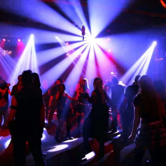 An image taken in a nightclub. You can see that people are dancing, but they are silhouetted against bright white lights and purple and red spotlights. Looks good!
