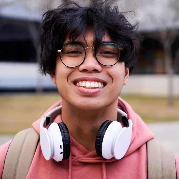 A photo of a student smiling widely. They are wearing round black-framed glasses, a pink hoodie, beige rucksack and he has white headphones around his neck.