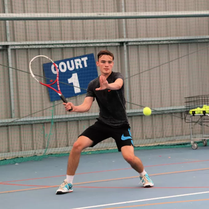 A photo of one of our English students in Eastbourne, on the tennis court, preparing to return the ball across the net. He is using his hand as a guide, and is focussing intently. In the background one of his coaches is looking on with interest. There is a blue sign behind him with white writing stating "Court 1".