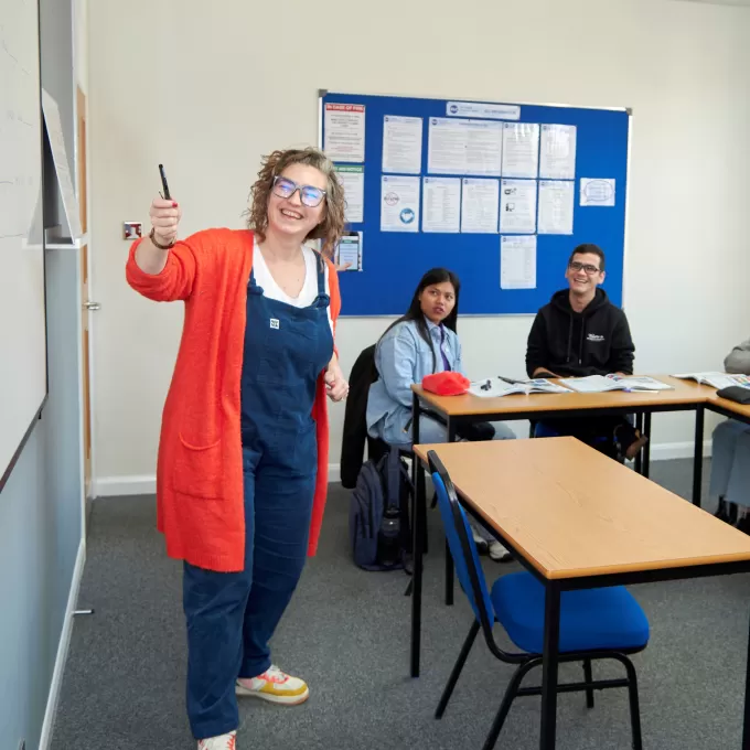 One of the teachers in Brighton pointing to the whiteboard in a photo that incorporates two of her students looking towards her. This photo is being used to highlight the teaching that happens in Brighton, and the teacher training courses for English teachers in Brighton.
