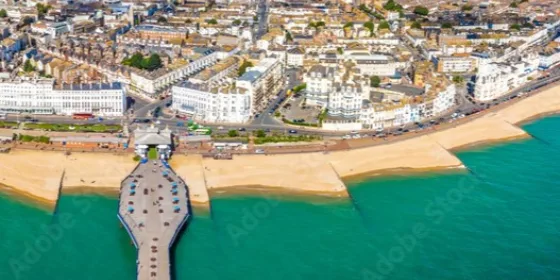 An aerial photo of Eastbourne's seafront, which uses a fish-eye lens to which makes it look like the beach is curving away from view. The pier is prominent in the shot, as is the sandy coloured beach. The sea is an inviting turquoise colour.