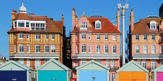 A view of five brightly painted Brighton beach huts which run along the promenade. Behind are three identical looking houses which look very grand, and which, today, will house a number of individual apartments. The sky in the background is very blue.