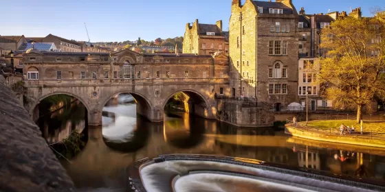 A photo of Pulteney Weir in Bath. It has a series of oval steps that the water runs over, which is visually appealing. In the background is Pulteney Bridge which is apparently one of only four bridges worldwide with shops across it on both sides.