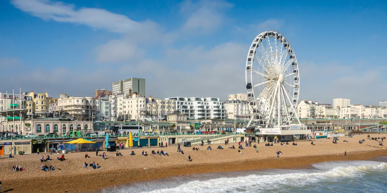 A photo of the seafront at Brighton which shows an array of white buildings and the sand-coloured pebble beach. There is a large white ferris wheel in the foreground, and a scattering of people sitting looking out to sea.