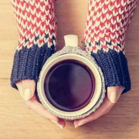 A close up shot of well manicured hands clutching a mug of black coffee. The wearer has a chunkily knitted jumper on with grey cuffs and orange and white sleeves, knitted in a chevron pattern.