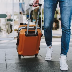 This is a photo of someone pulling a new-looking, bright orange wheeled suitcase along the pavement. You can only see their bottom half. They are wearing blue jeans and white converse-style trainers.
