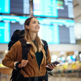 A photo suggestive of an English language student with her backpack staring up at a screen. In the background, quite blurred are big departure boards that you would find in an airport. She has her passport and tickets in her hand.