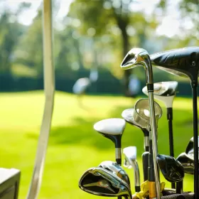 A photo of the heads of golf clubs in their golf bag. The background is blurred, but you can clearly see a putting green and someone taking a putt. This is representative of what you might see on the English language plus golf option of ELC Eastbourne's English and activity programme.