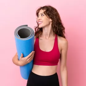 A library shot of someone who looks like they're about to do some yoga. She's carrying a rolled up yoga mat, looking off the her right and the photo is taken against a pink background.