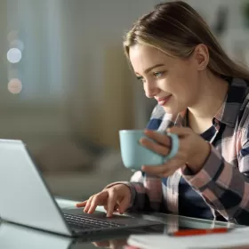 A student sits at a desk, her hand on her laptop. She's looking at the screen and smiling whilst holding a blue cup. Let's hope she doesn't spill tea on her keyboard.