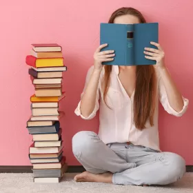 A woman's face is hidden by the hardback book she's reading. It's very close to her eyes. There's a big pile of other books next to her, and she's sitting crosslegged, her back against a pink wall. This is clearly all about reading.