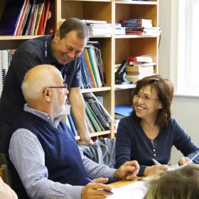 A photo of two over 50s English course students in Chester chatting to their teacher. Behind them is a bookshelf suggesting they may be doing some work in the library.