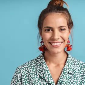A close up head shot of a woman with her hair tied back revealing a pair of red, dangly earrings. She has a great and white blouse and she's photographed against a plain blue background.