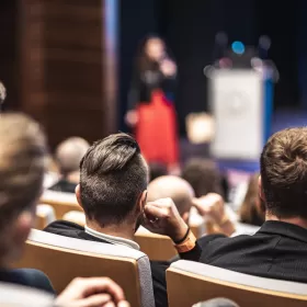 This image is taken from a lecture hall where a lecturer is standing on stage, very out of focus, but clearly wearing a red skirt and black jacket. The students look like they might be business students and are sitting in raked seating in the auditorium.