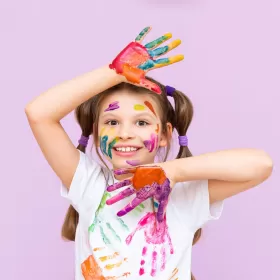 A library photo of a young girl who has clearly been having a good time with a load of paints. Her open palms face the camera and they're covered with different paint colours. She's got hand prints on her white T-shirt and more paint on her face. The background is a muted pink colour.