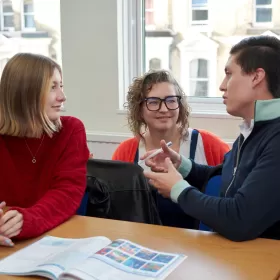 A photo of English language students Elisa Hentsch and Jorge Filipe Suesca Romero talking to each other during their English course in Brighton. The teacher is monitoring their conversation with a big smile on her face. The students must be doing well.